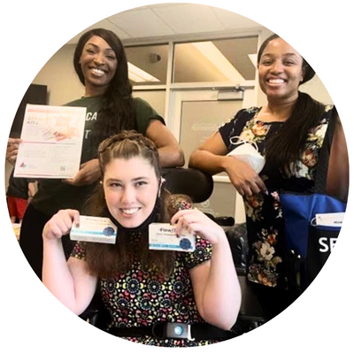 Three women, two standing and using a manual wheelchair holding up bags with covid tests,